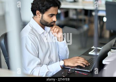 Busy young indian employee working on laptop, looking at computer in office. Stock Photo