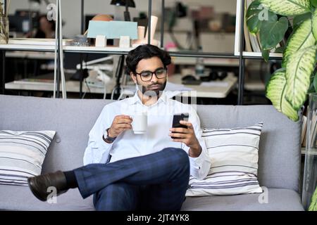 Indian business man using mobile phone reading news drinking coffee in office. Stock Photo