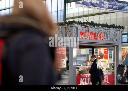 Stuttgart, Germany - December 31, 2021: Man with corona mouth and nose mask, shopping in butcher shop. Snack bar in main station hall is decorated for Stock Photo