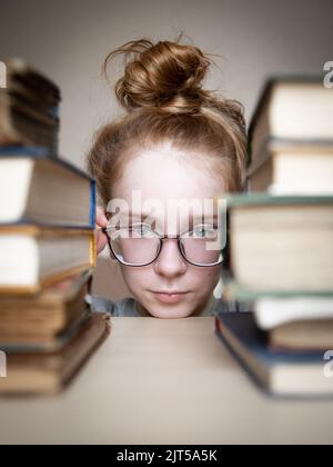 Beautiful teenage girl with red hair tied in bun looks between two stacks of hardback books lying on light table. Girl holding glasses on her face Stock Photo