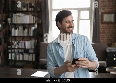 Cheerful dreamy millennial smartphone user man holding cell Stock Photo