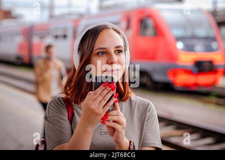 Smiling woman listens to music with headphones using smartphone on railway platform. Stock Photo