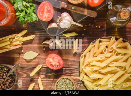 Raw penne pasta bowl placed with fresh ingredients on rustic wooden background. Top view with close up. Stock Photo