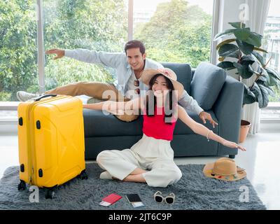 Happy Asian couple lover ready to trip, young woman and man in colorful casual making gesturing airplane wings, smiling together, sit on floor with pa Stock Photo