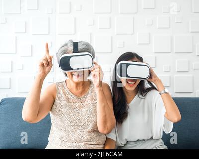 Asian young woman, happy daughter and her mother, old senior female wearing VR glasses, enjoy 3d game online together in white room. Mom and daughter Stock Photo