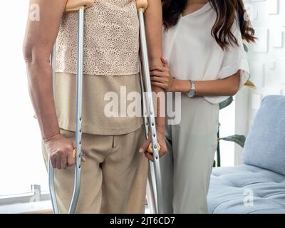 Person helping, female's hand hold the arms of an elderly woman trying to walk on crutches standing in recovery room background. Helping and care seni Stock Photo