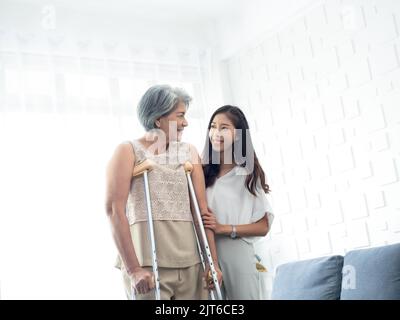 Young smiling Asian females carefully hold and support the arms of elderly women trying to walk on crutches in recovery room, helping old women, healt Stock Photo