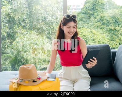 Young beautiful happy Asian woman with sunglasses using digital tablet computer with passport, flight ticket and beach hat on yellow suitcase, sit on Stock Photo