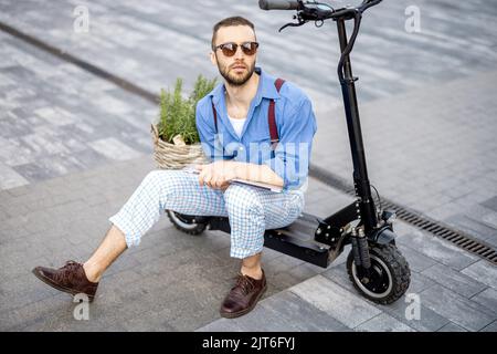 Portrait of stylish weird man sit with laptop on electric scooter outdoors Stock Photo