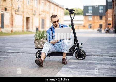 Man works on laptop while sitting on electric scooter outdoors Stock Photo