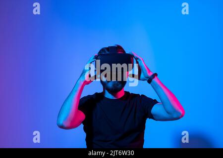 Young bearded man playing video games in VR-glasses, experiencing virtual reality while standing in neon light over blue gradient background. Stock Photo