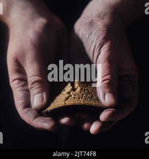 Hungry man holding bread on a black background, hands with food close-up. Dirty hands of a starving poor man on a dark background Stock Photo