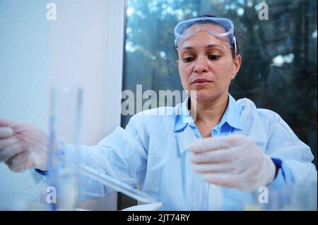 Multi-ethnic experienced female chemist scientist in lab coat using labware while working in medical research laboratory Stock Photo