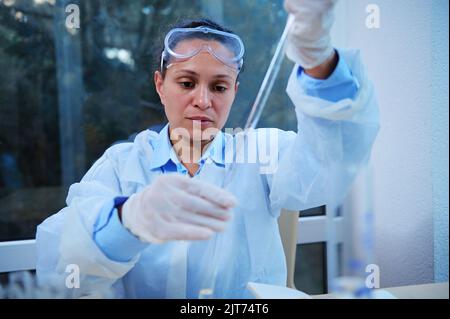 A female laboratory assistant using graduated pipette drips a clear liquid into a test tube with a special preparation. Stock Photo