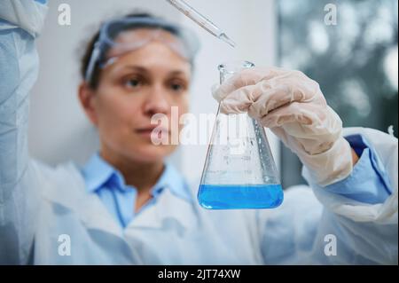 Focus on flat-bottomed flask and blurred charming woman scientist dripping few reagent into blue solution with a pipette Stock Photo