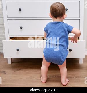 Toddler baby opens a chest of drawers. Child boy reaches into an open drawer of a white cabinet. Kid age one year Stock Photo