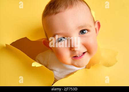 A happy child looks out of a hole in the studio yellow background. Smiling baby boy peeks through a torn paper background, copy space. Kid age one yea Stock Photo