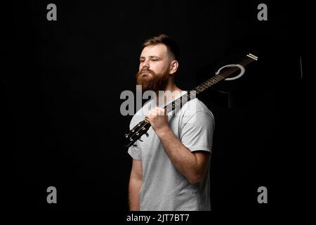 Young bearded guitarist in grey t-shirt holds a guitar on a shoulder in a dark room. Stock Photo