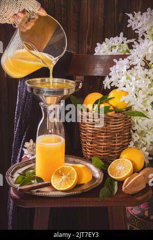 Pouring fresh orange juice from jug into glass bottle. Still life with orange juice bottle placed with sliced oranges and wooden squeezer. Stock Photo
