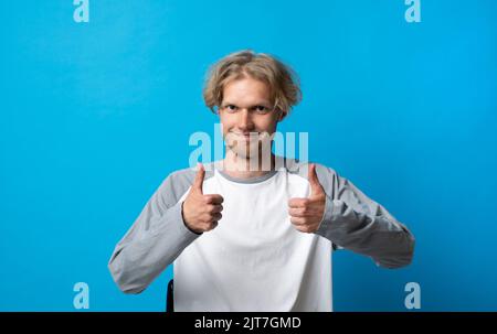 Young handsome blonde man in white t-shirt doing happy thumbs up gesture with both hand. Approving expression looking at the camera, showing success. Stock Photo
