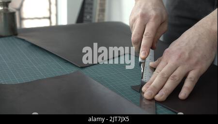 Working process of crafting a new handmade leather wallet in the leather workshop. Man's hands working with knife and iron ruler. Stock Photo