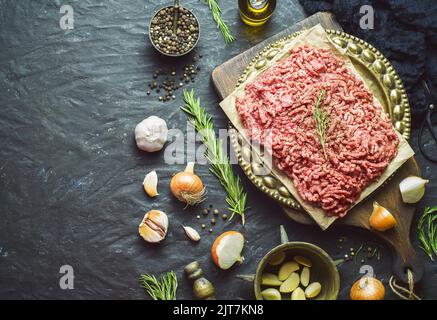 Fresh raw minced beef place with peppercorn, virgin olive oil, garlic and onion on dark background. Top view with copy space. Stock Photo