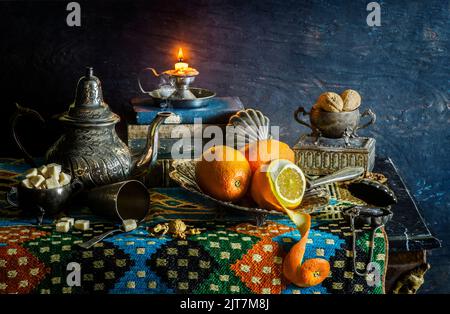 Classic still life with fresh oranges in silver plate placed with illuminated candle, old books, nuts, sugar, silver kettle on ornate cloth.. Stock Photo