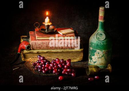 Classic still life with fresh delicious grapes placed with vintage wine bottle, old books, pipe and illuminated candle on rustic dark background.. Stock Photo