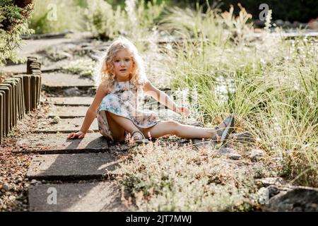 Portrait of poor crying awesome little girl with long fluffy curly hair wearing dress with stars, sitting after falling. Stock Photo