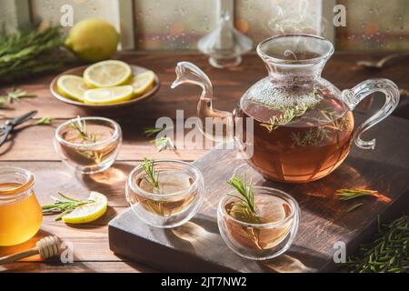 Hot rosemary tea in glass teapot and cups. Served with honey and fresh lemon on rustic wooden background. Stock Photo