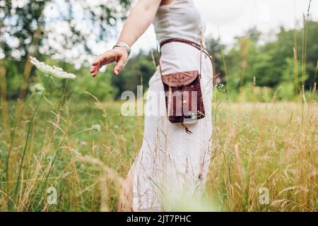 Boho style fashion. Woman wearing white dress, jewelry, carrying leather handmade handbag outdoors walking in meadow Stock Photo