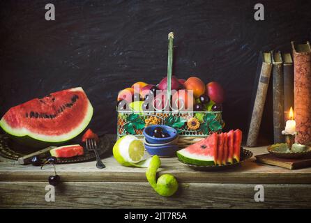 Classic still life with various fruits placed in vintage basket,fresh watermelon slices, old books and illuminated candle on rustic wooden background. Stock Photo