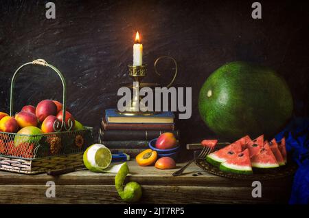 Classic still life with various fruits placed in vintage basket, watermelon slices, old books and illuminated candle on rustic wooden background. Stock Photo