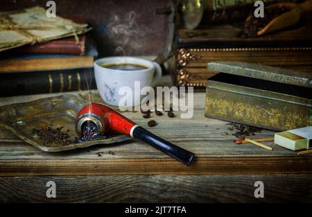 Classic still life with vintage pipe placed with tobacco, cup of coffee, old books, coffee grinder and coffee beans on rustic wooden background. Stock Photo