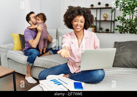 Mother of interracial family working using computer laptop at home smiling cheerful offering palm hand giving assistance and acceptance. Stock Photo