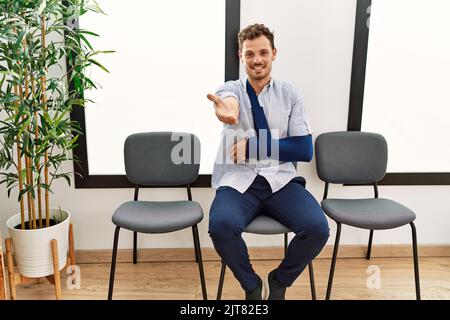 Handsome young man sitting at doctor waiting room with arm injury smiling cheerful offering palm hand giving assistance and acceptance. Stock Photo