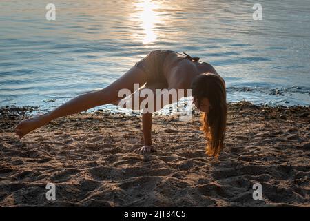 Young beautiful slim woman is doing yoga on the seashore on the sand Stock Photo