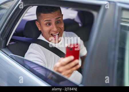 Young hispanic man doing video call with smartphone in the car celebrating achievement with happy smile and winner expression with raised hand Stock Photo