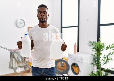 Young african man doing laundry holding detergent bottles making fish face with mouth and squinting eyes, crazy and comical. Stock Photo
