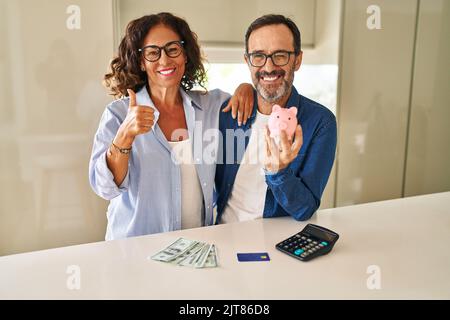 Middle age couple holding piggy bank calculating savings smiling happy and positive, thumb up doing excellent and approval sign Stock Photo