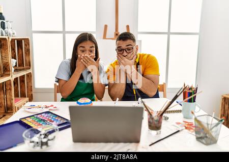 Young hispanic couple using laptop sitting on the table at art studio shocked covering mouth with hands for mistake. secret concept. Stock Photo