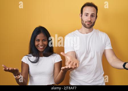 Interracial couple standing over yellow background smiling cheerful offering hands giving assistance and acceptance. Stock Photo