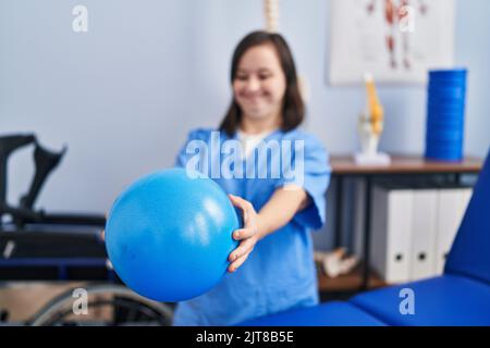 Down syndrome woman wearing physiotherapy uniform holding ball at physiotherapist clinic Stock Photo