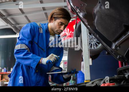 Young man is mechanic using laptop computer checking engine of car in the garage, worker examining and diagnostic automobile, technician and maintenan Stock Photo