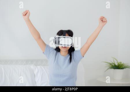 Young asian woman wearing vr headset playing game with excited in the bedroom at home, female using virtual reality or metaverse innovation for simula Stock Photo