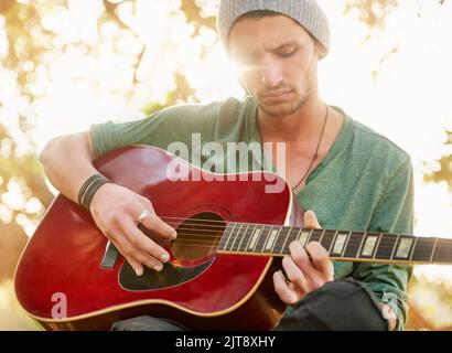 Nature has the best acoustics. a handsome young man sitting outside in a park playing guitar. Stock Photo