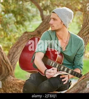 Getting musically inspired by nature. a handsome young man sitting in a park with a guitar writing song on a notepad. Stock Photo