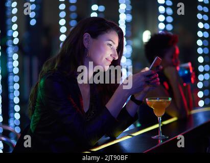 When are you getting to the club. a smiling young woman reading a text message while sitting at the bar in a nightclub. Stock Photo