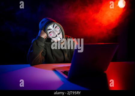 June 5, 2022 Novosibirsk, Russia: Anonymous in a hood is typing on a laptop in the dark in red-blue smoke.  Stock Photo