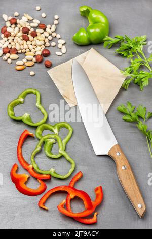 Chopped green and red pepper. Kitchen knife on paper, nuts and chickpeas, parsley sprigs on table. Flat lay. Grey background. Stock Photo